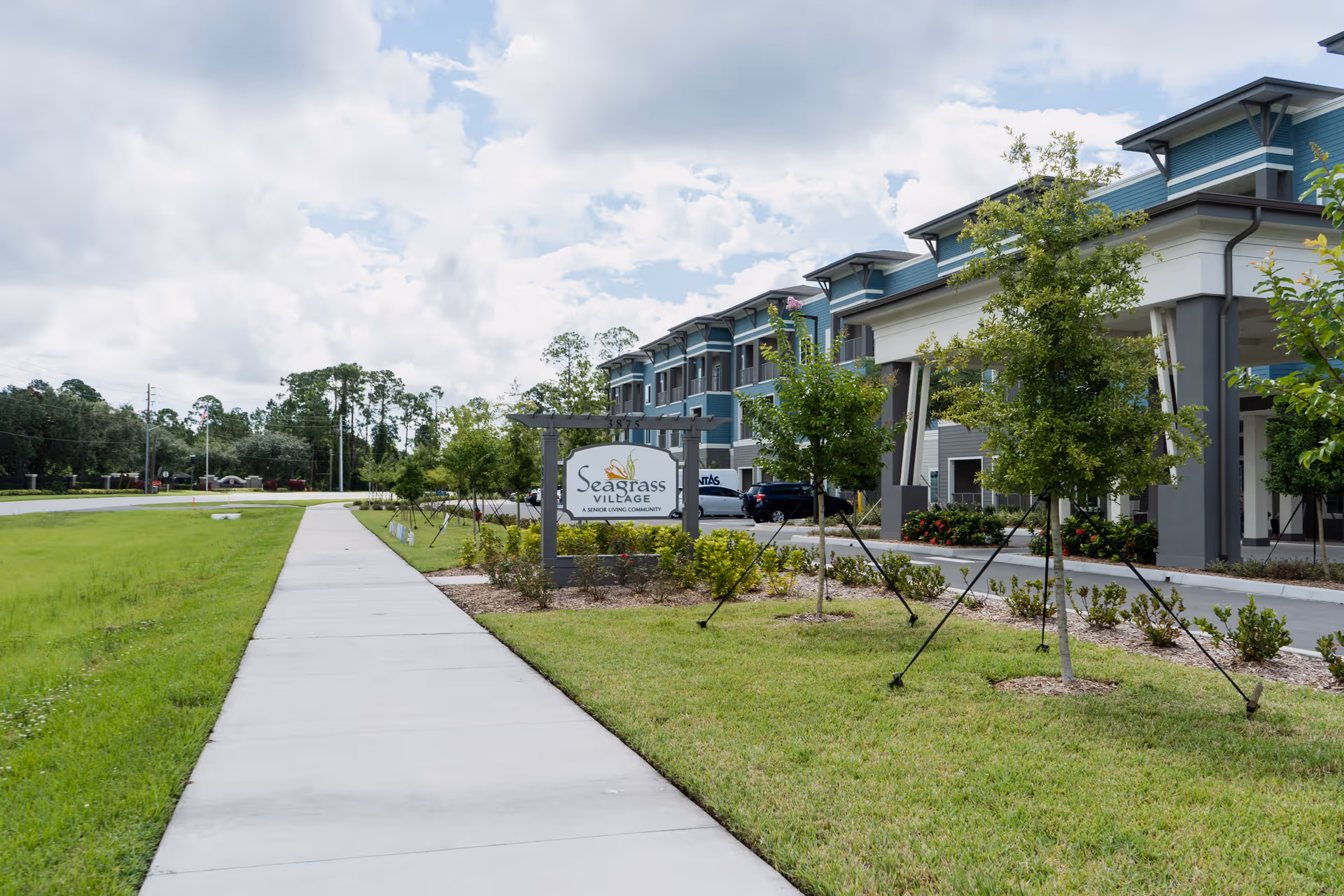Sidewalk leading to the entrance of Seagrass Village of Port Orange, a senior living community, with a landscaped lawn and young trees supported by stakes. The building is modern with blue and gray exterior walls and multiple windows under a partly cloudy sky.