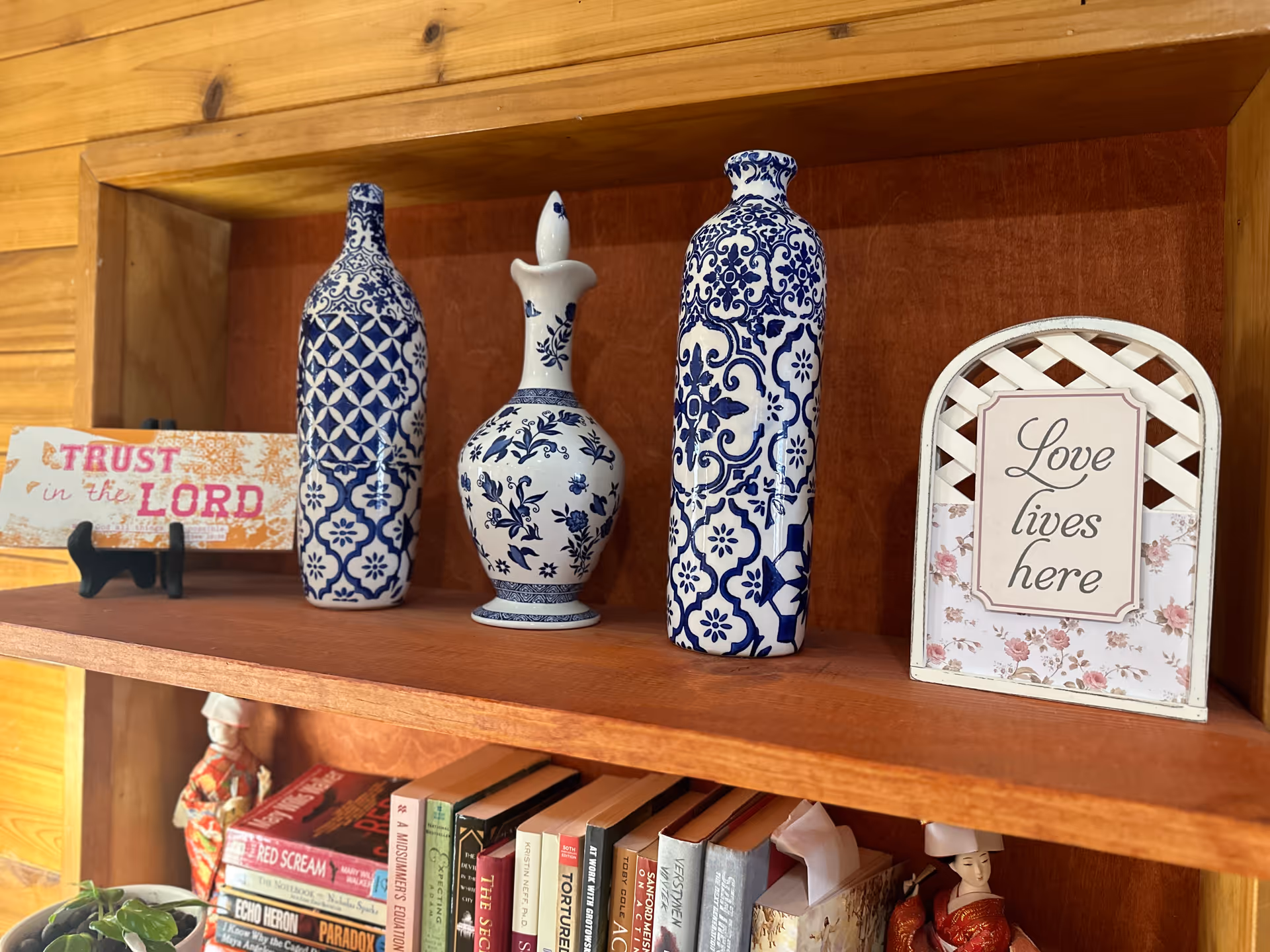 Wooden shelf displaying three blue and white decorative vases, a sign that reads 'Trust in the Lord,' and another sign that says 'Love lives here.' Below the shelf are books and small decorative items including a plant and figurines.