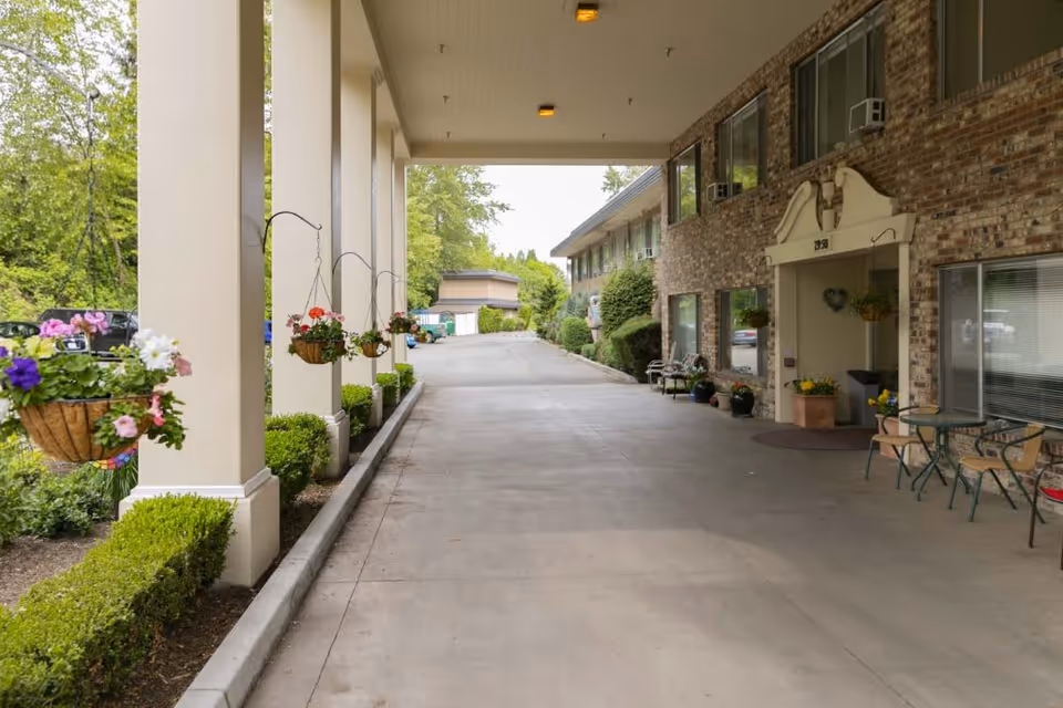 Covered driveway entrance to a senior living facility with hanging flower baskets on columns, trimmed bushes along the side, and a brick building with windows and a small seating area with chairs and a table near the entrance.