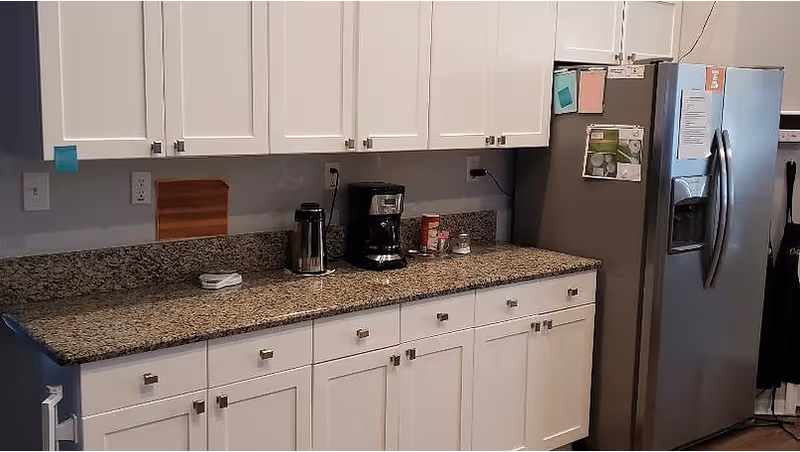 A kitchen area with white cabinets and drawers, a granite countertop with a coffee maker, a kettle, and some small containers. A stainless steel refrigerator is on the right side with notes and pictures attached to it.