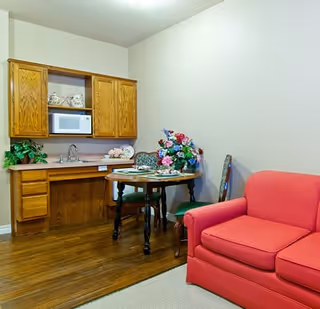 Small living area with a red loveseat, a round table topped with a floral centerpiece, and a kitchenette with wooden cabinets and a microwave.