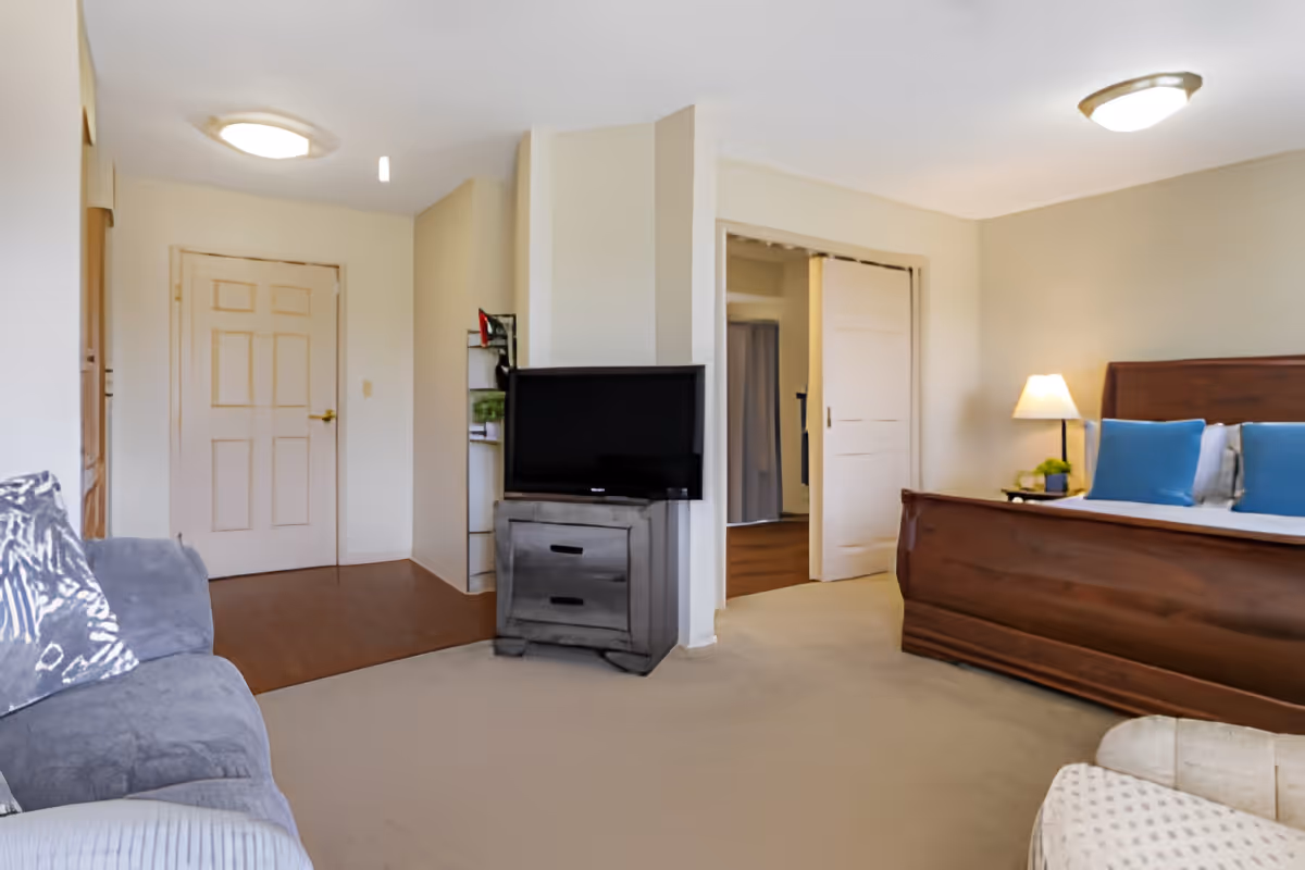 A cozy senior living bedroom featuring a wooden bed with blue pillows, a bedside table with a lamp and plant, a TV on a small wooden dresser, a gray couch with patterned pillows, and a beige carpeted floor. The room has cream-colored walls and two ceiling lights, with a door and an open sliding door leading to other areas.