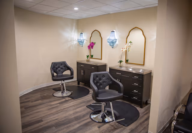 Interior view of a small salon area with two black salon chairs in front of two dark wood cabinets with mirrors above them. Each cabinet has a vase with flowers, and there are wall-mounted light fixtures above the mirrors. The room has light-colored walls and wood flooring.