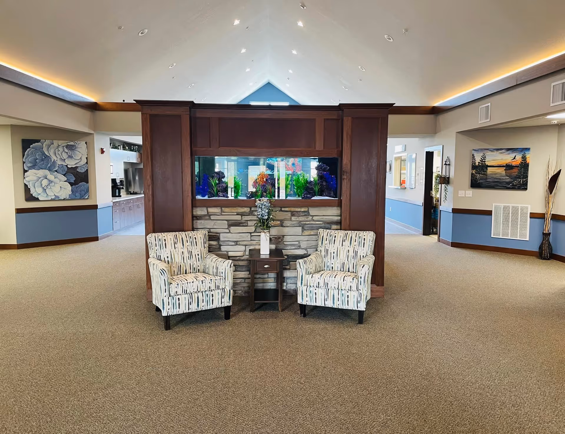Spacious senior living common area with two patterned armchairs and a small table in front of a built-in aquarium beneath a vaulted ceiling.