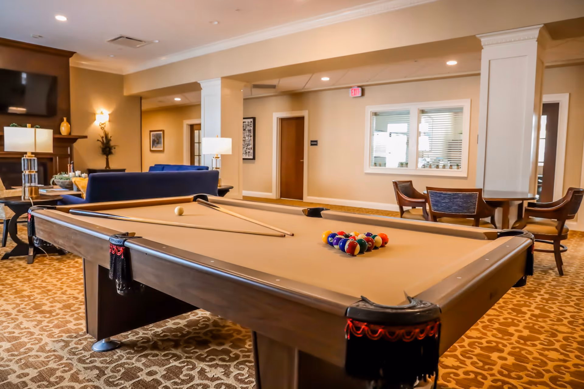 A spacious common area featuring a pool table with billiard balls and cues set up, surrounded by comfortable seating including blue sofas and wooden chairs. The room has warm lighting, a patterned carpet, and a wall-mounted TV above a fireplace.