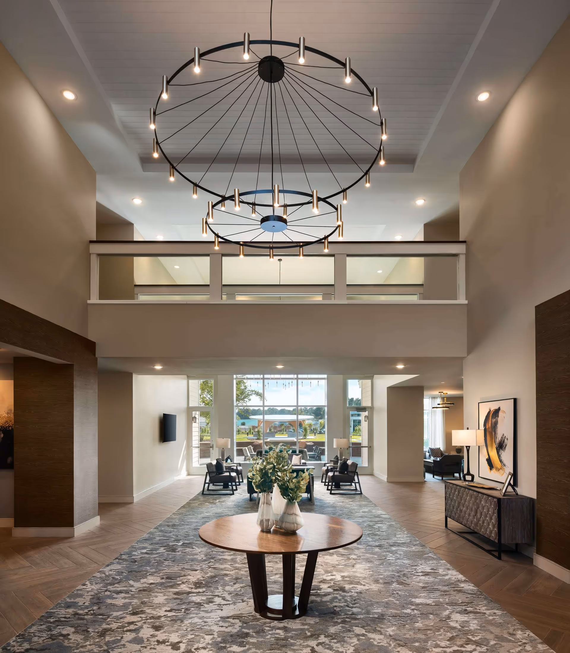 Spacious and modern interior of a senior living facility lobby with high ceilings, a large circular chandelier, a round wooden table with vases of flowers in the center, seating areas with chairs and lamps near large windows letting in natural light, and contemporary artwork on the walls.