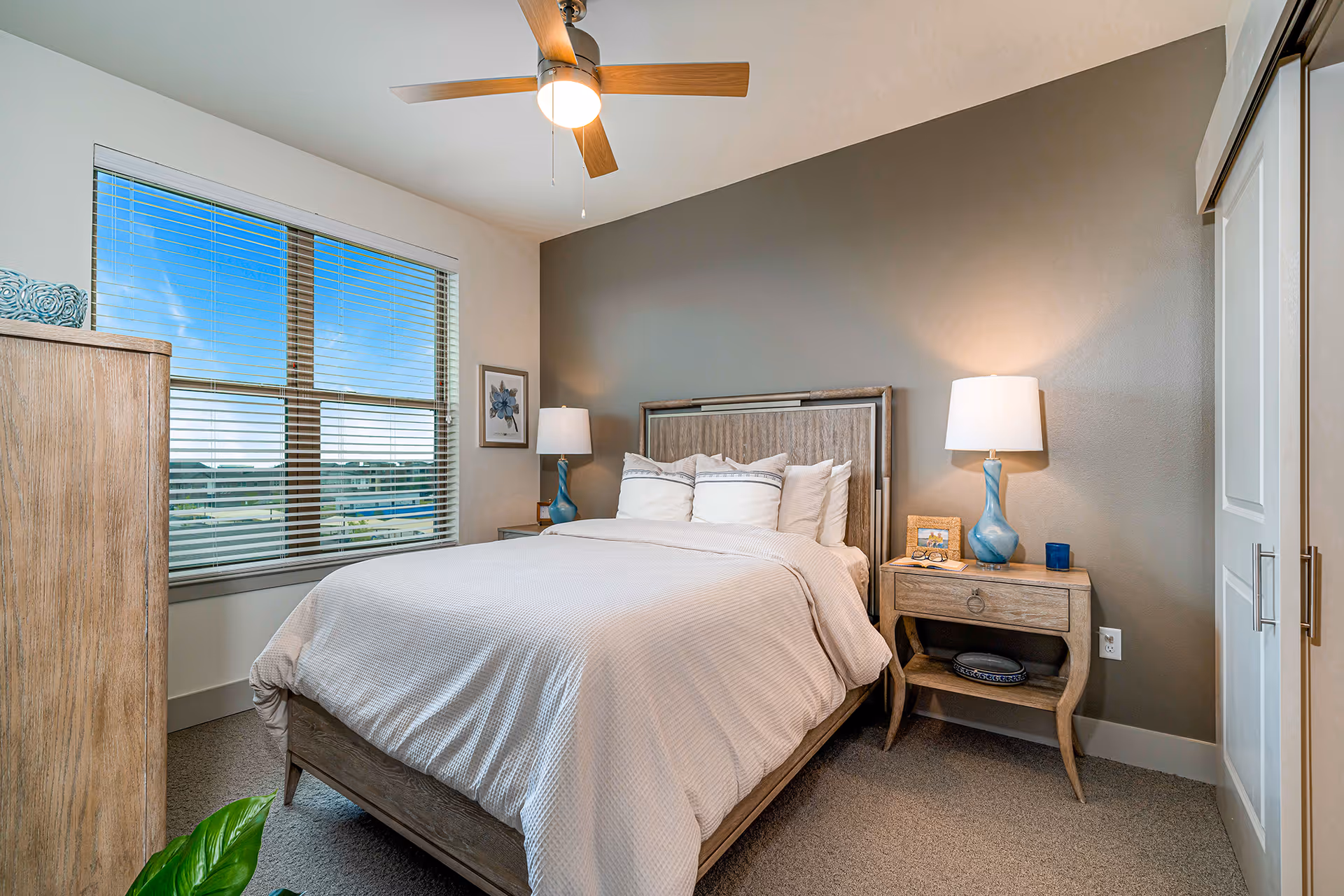 A cozy bedroom with a large bed featuring white bedding and multiple pillows. The room has a wooden headboard and matching wooden nightstands on each side, each with a blue lamp. A window with blinds lets in natural light, and a ceiling fan with a light is mounted above the bed. The walls are painted in neutral tones, with one accent wall in gray. There is a framed floral picture on the wall next to the window and a closet with sliding doors on the right side.