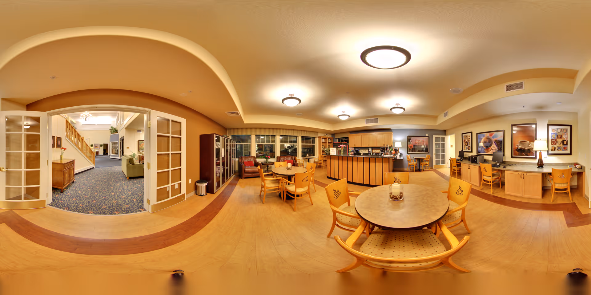 A spacious and well-lit common area in a senior living facility featuring multiple round tables with chairs, a small kitchen or coffee bar area with cabinets and appliances, and a seating area with armchairs near large windows. The room has warm yellow walls, wood flooring with a darker wood border, and ceiling lights. There is a hallway visible through glass double doors leading to another part of the facility with carpeted flooring and a staircase.