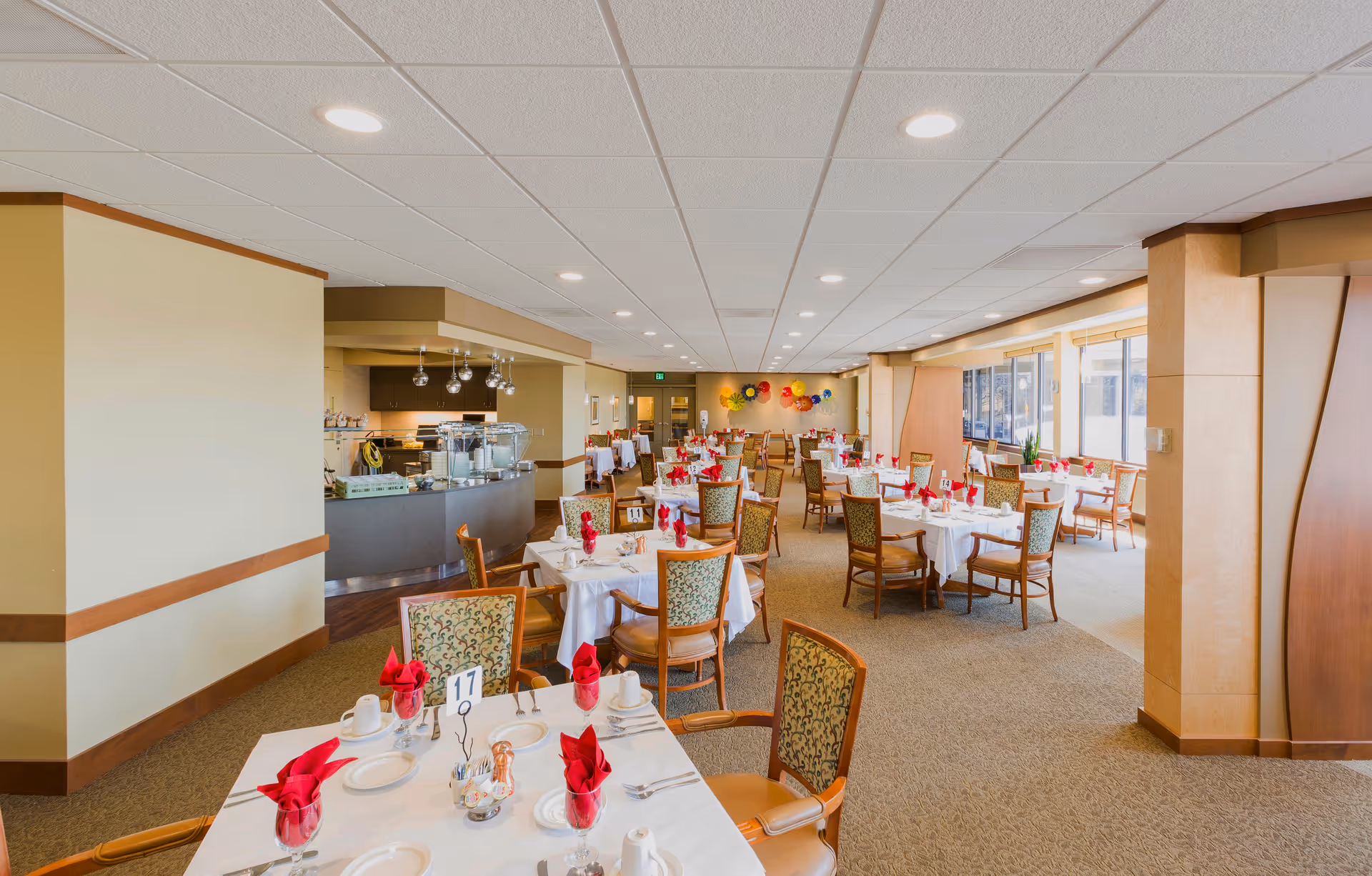 A spacious dining room in a senior living facility with multiple tables set with white tablecloths, red folded napkins, plates, cups, and silverware. The room has patterned chairs, large windows letting in natural light, and a serving area with hanging lights. Colorful wall decorations are visible at the far end of the room.