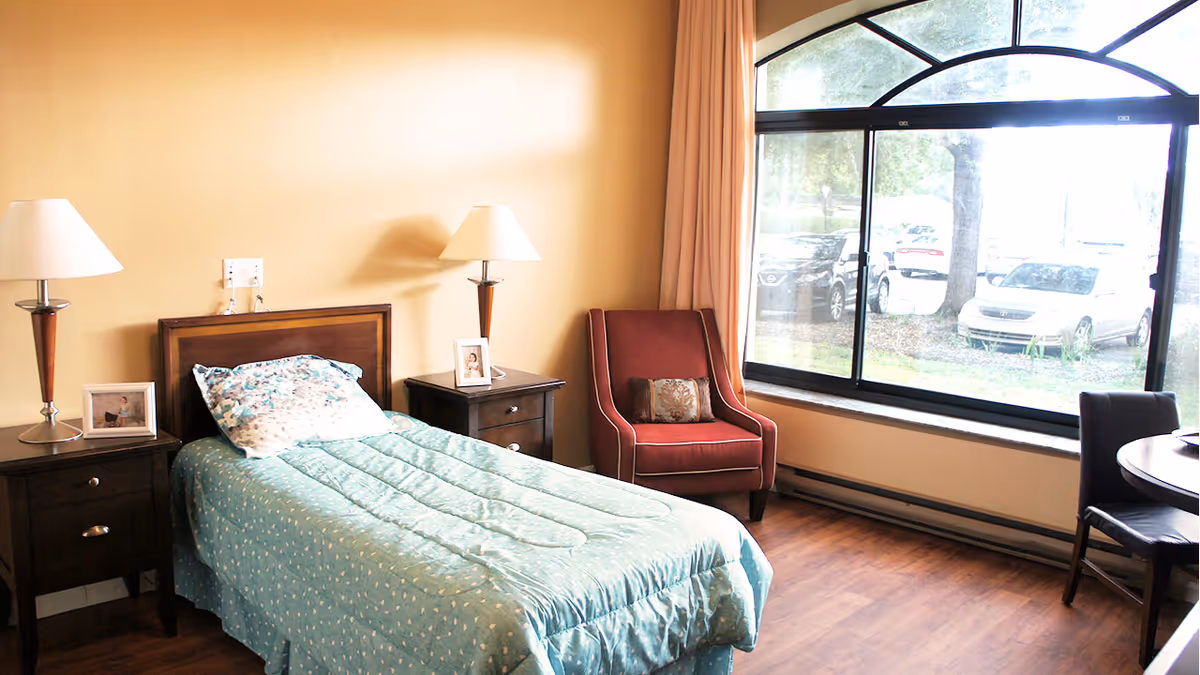 A cozy bedroom with a single bed covered in a light blue patterned comforter and a matching pillow. The bed is flanked by two dark wooden nightstands, each with a lamp and a framed photo. A red armchair with a decorative pillow is positioned near a large window that lets in natural light and offers a view of parked cars and trees outside. The room has wooden flooring and beige walls.