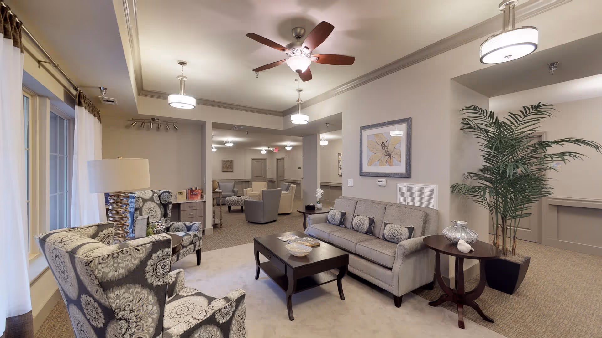 A well-lit living room area in a senior living facility featuring a gray sofa with patterned cushions, two matching armchairs with floral designs, a dark wooden coffee table with decorative bowls, a side table with a lamp, a large potted plant, ceiling fans with lights, and framed artwork on the wall.