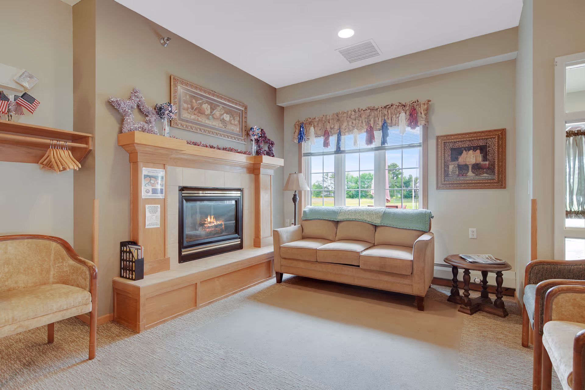 A cozy living room with a beige sofa, a wooden fireplace with a fire burning, a window with decorative curtains, a small wooden side table with magazines, and two armchairs. The room has warm beige walls and carpeted flooring.