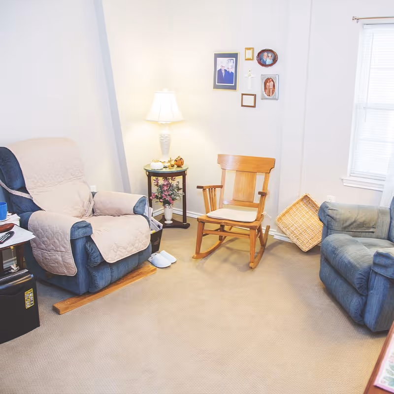 A cozy living room with a blue recliner covered by a beige quilted cover, a wooden rocking chair with a cushion, a small round side table with a lamp and decorative items, family photos on the wall, a window with blinds, and a wicker basket leaning against the wall.