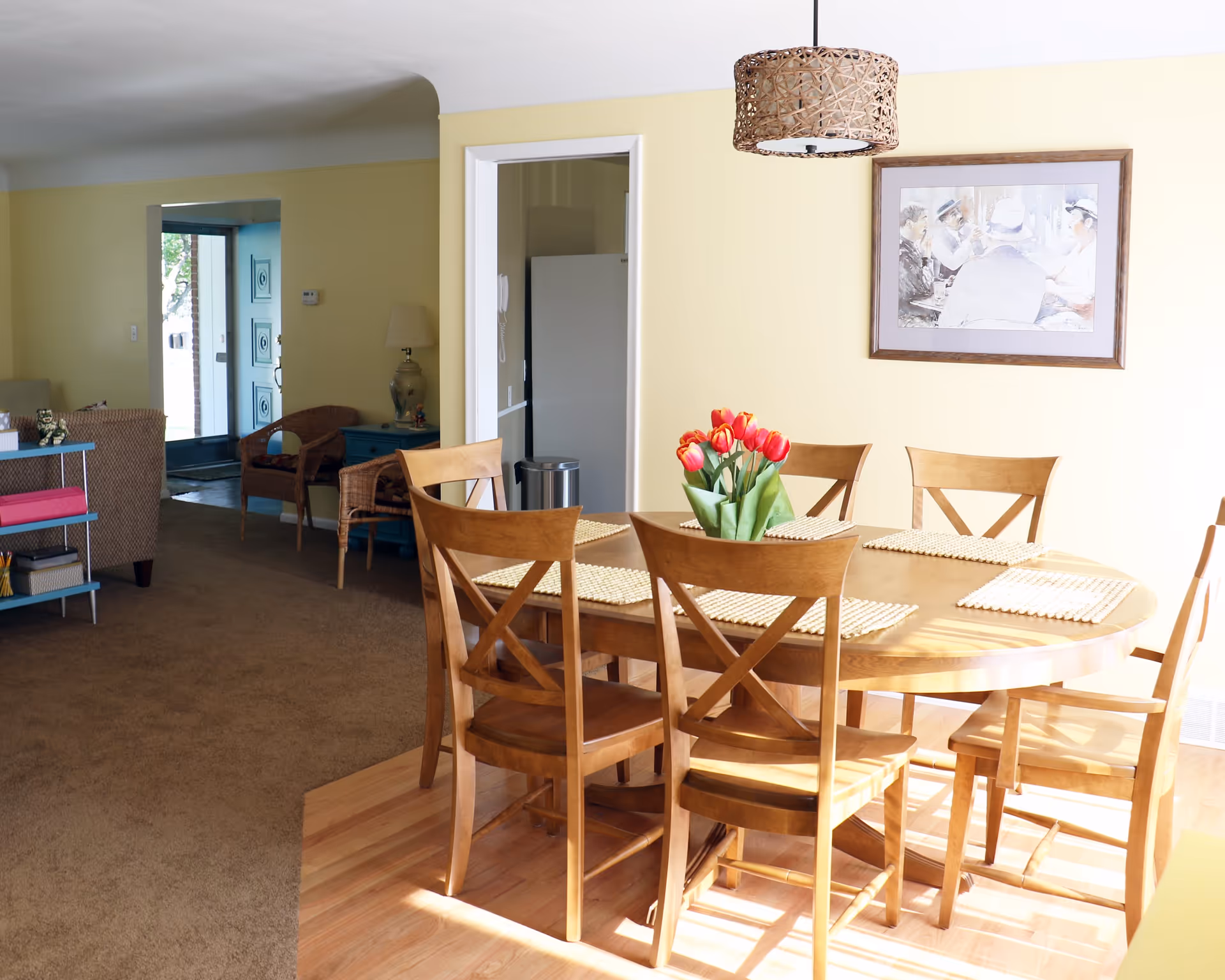 A bright dining area with a wooden oval table surrounded by six wooden chairs. A vase with red tulips is placed in the center of the table on woven placemats. The room has light yellow walls, a wicker pendant light hanging from the ceiling, and a framed painting on the wall. In the background, there is a view into a living room area with wicker chairs, a blue side table with a lamp, and an open front door letting in natural light.