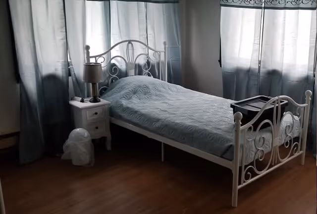 A simple bedroom with a single white metal bed frame, a light blue quilted bedspread, a white nightstand with a lamp, and two windows with light blue curtains allowing natural light to enter the room.