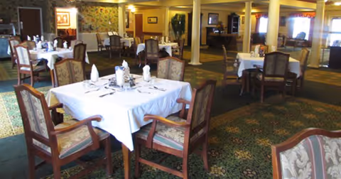 Dining room with multiple tables draped in white tablecloths, wooden chairs, carpeted floor, and supporting columns.