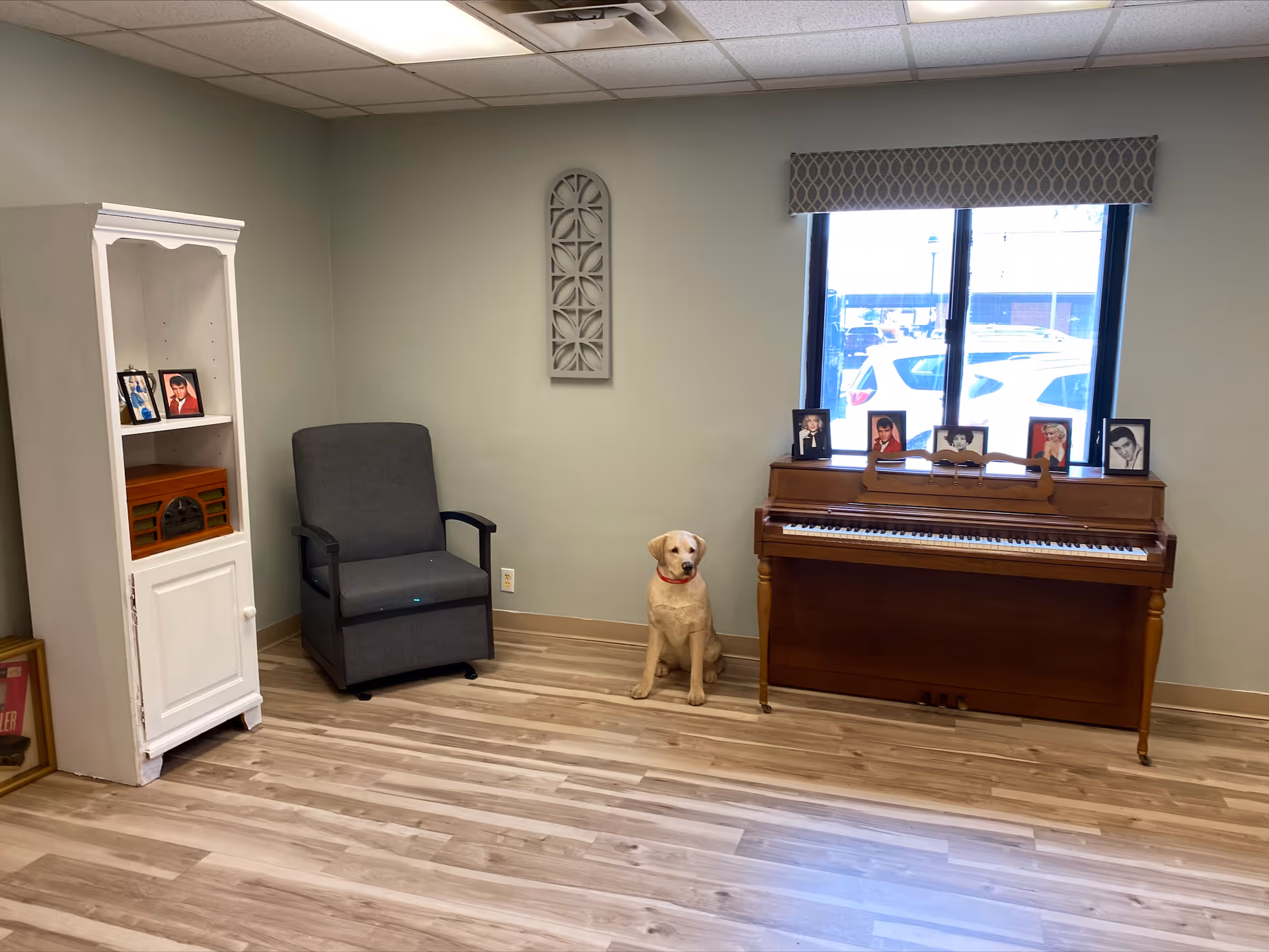 Small common room with a piano, armchair, white shelving unit and a life-size dog statue by a window.