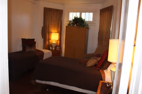 A cozy bedroom with two beds covered in brown bedding, a wooden nightstand with a lamp, a wooden cabinet with a plant on top, and two windows with decorative curtains.