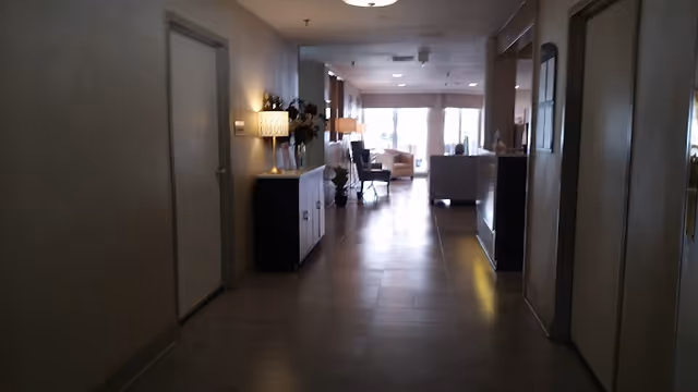 A dimly lit hallway in a senior living facility with closed doors on both sides, a small cabinet with a lamp and flowers on the left, and a seating area with chairs and large windows at the far end.