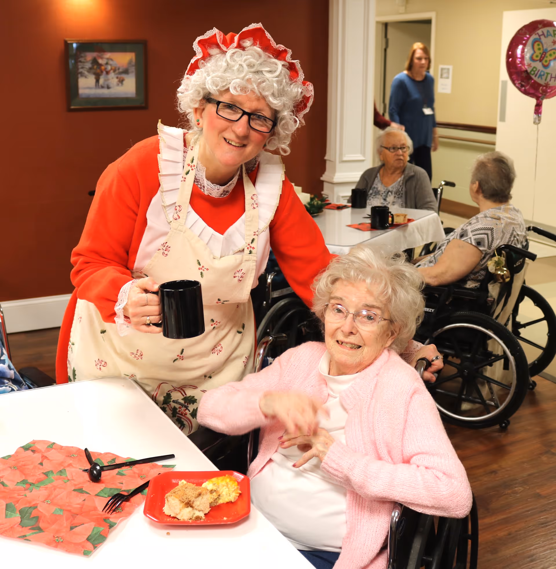 A woman in a festive apron and wig holds a mug while smiling next to a seated elderly woman in a wheelchair at a dining table in a care facility.