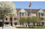 Exterior view of a two-story brick building with multiple windows and green awnings. There is a flagpole with an American flag in front of the building, surrounded by neatly trimmed trees and landscaping under a clear blue sky.