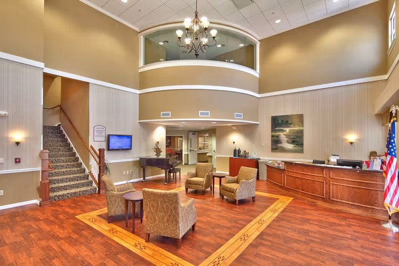 Spacious senior living facility lobby with wooden flooring and a decorative inlay. Four patterned armchairs arranged around two small round tables in the center. A grand piano is placed near the back wall. Reception desk with a staff member seated behind it is on the right side. An American flag stands next to the reception desk. A staircase with carpeted steps and wooden railing is on the left side. Walls are painted beige with white trim, and a chandelier hangs from the high ceiling. A large painting is mounted on the wall behind the reception desk.