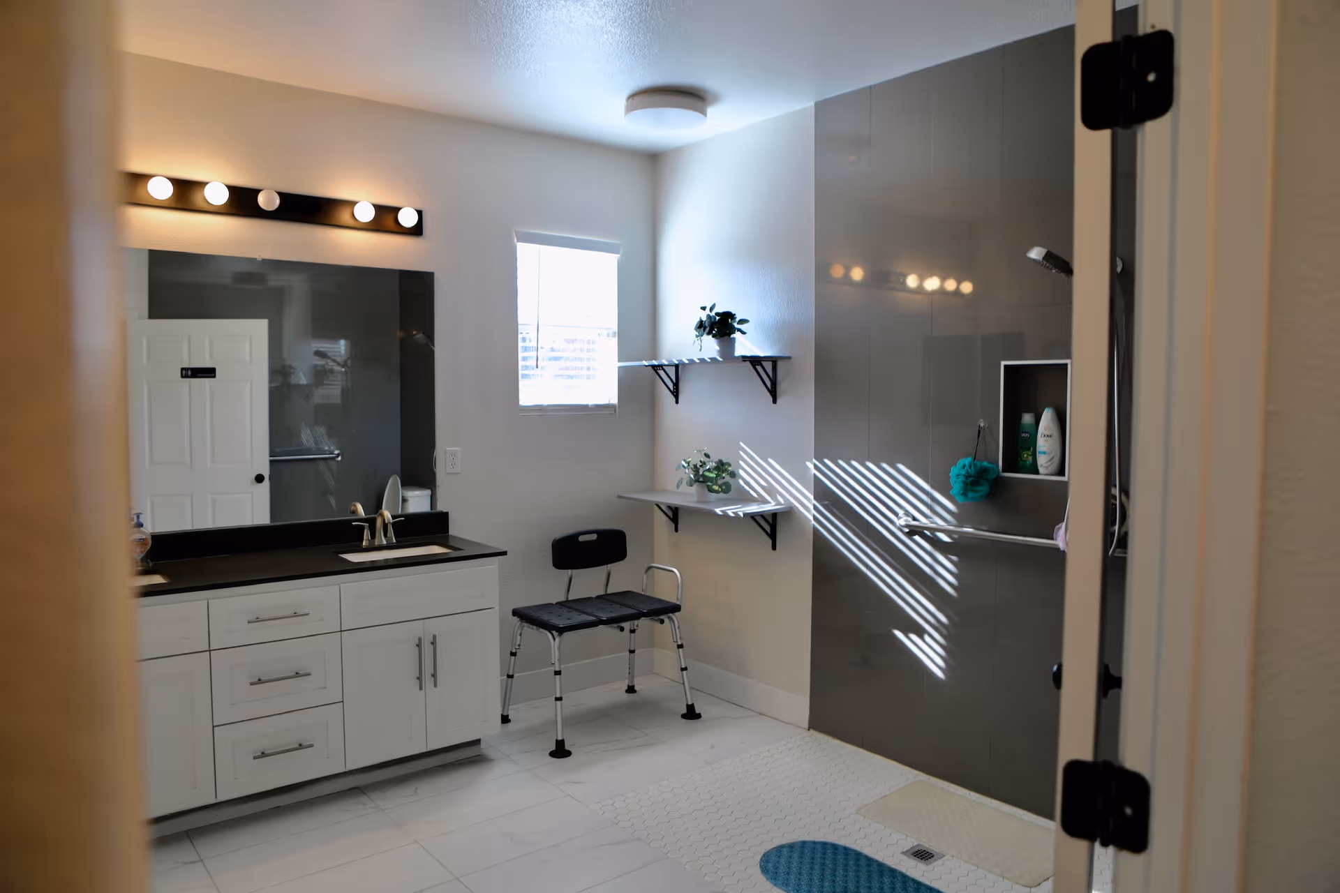 A bright bathroom with a white vanity and black countertop, a large mirror with lights above it, a small window letting in natural light, two wall-mounted shelves with plants, a black shower chair, and a walk-in shower area with a grab bar and toiletries.