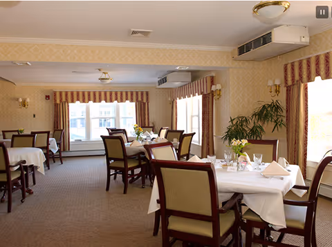 A dining room with several tables covered in white tablecloths, each set with napkins, glasses, and small floral centerpieces. The room has beige patterned wallpaper, large windows with striped curtains, and upholstered chairs around the tables. Ceiling lights and wall sconces provide lighting.