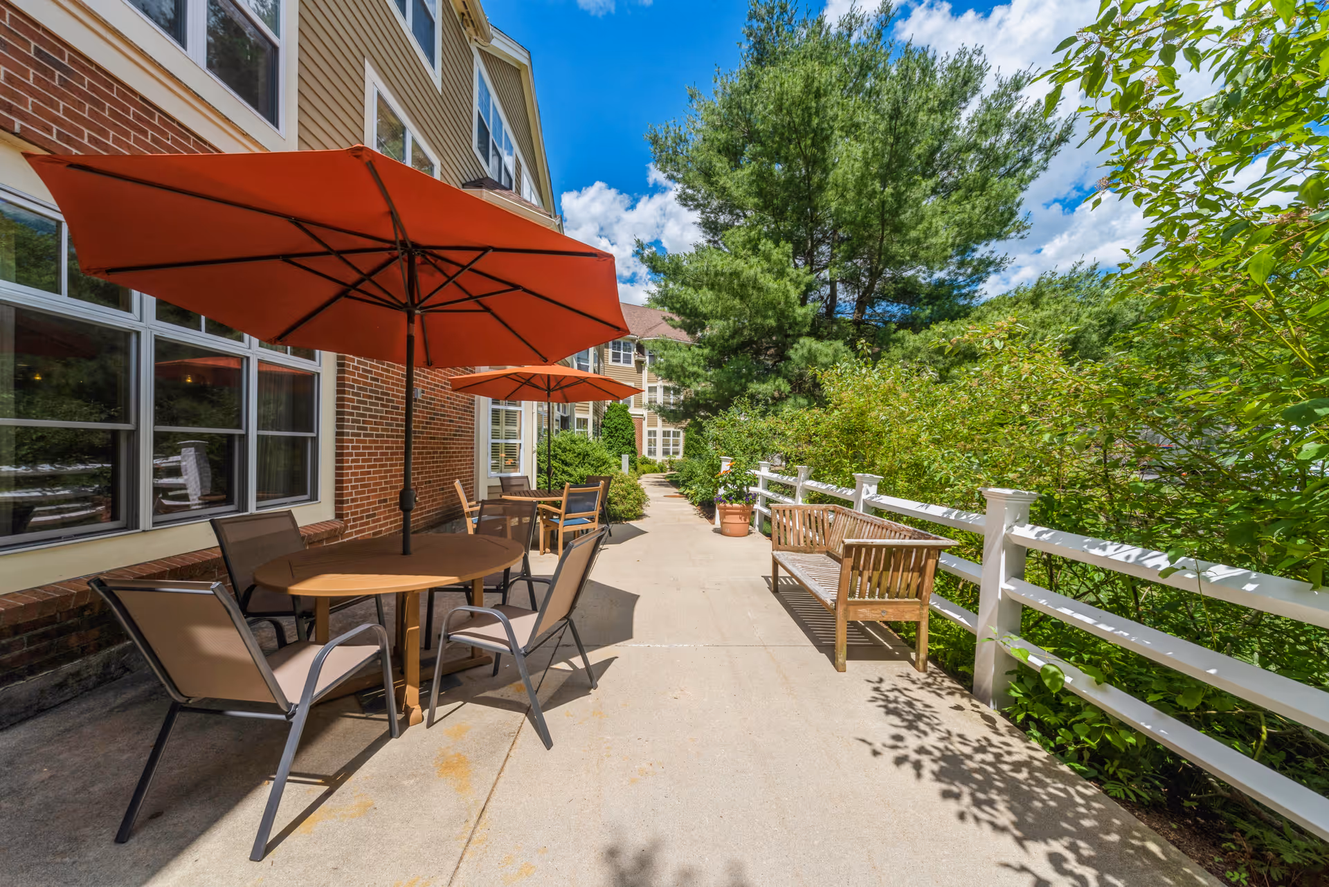 A sunny outdoor patio with round tables, red umbrellas, chairs and wooden benches along a building and white fence next to green landscaping.