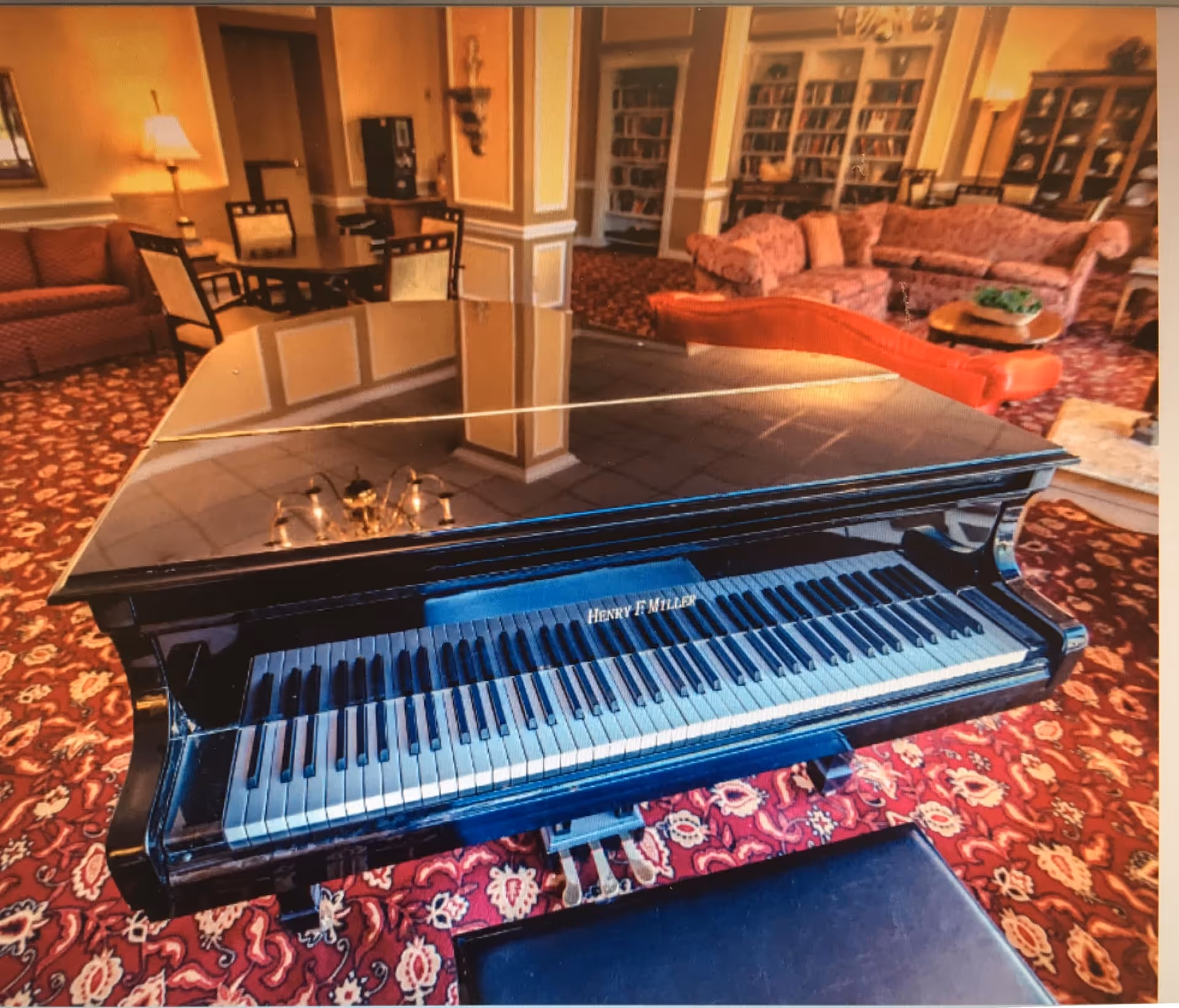 A black grand piano with the name Henry F Miller on it is placed in a cozy living room area with patterned red carpet. The room features multiple seating arrangements including sofas and chairs, bookshelves filled with books, and warm lighting from lamps and chandeliers.