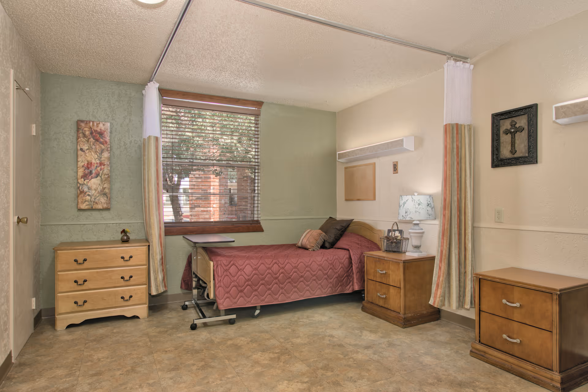 A simple, clean bedroom in a senior living facility with a single bed covered in a maroon quilt. The room has a window with blinds and curtains, a small wooden dresser, a bedside table with a lamp, and another small wooden cabinet. There is a decorative cross on the wall and a floral painting near the door.