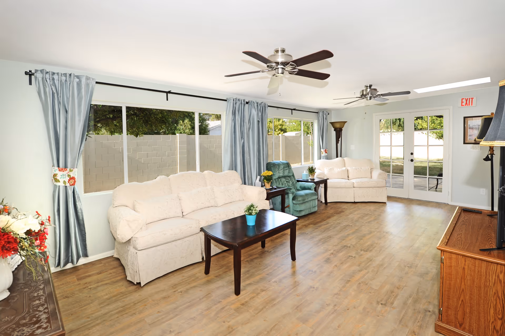 A bright living room with two cream-colored sofas, a green recliner chair, a dark wooden coffee table with a small potted plant, and large windows with light blue curtains. The room has wooden flooring, two ceiling fans, a floor lamp, and a glass door leading outside with an exit sign above it.