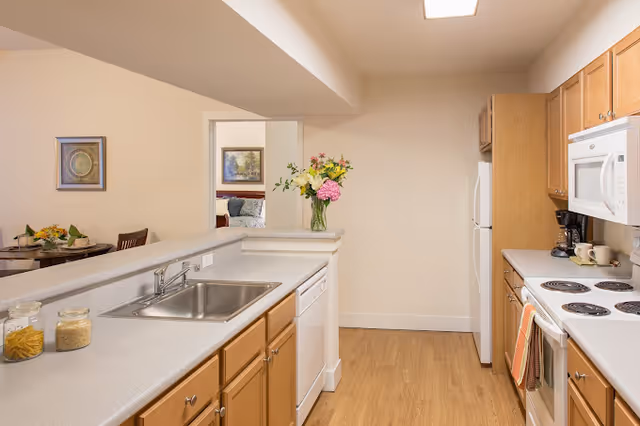 Galley-style kitchen with light wood cabinets, white appliances, a sink and dishwasher, and a vase of flowers on the counter.
