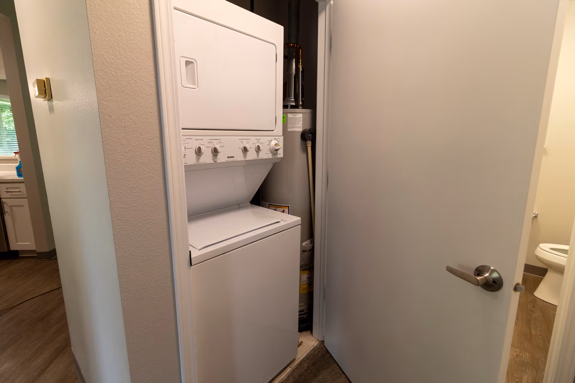 A small laundry area with a stacked washer and dryer unit next to a water heater, partially visible behind an open door. To the right, a bathroom with a toilet is visible through another open door. The floor is wood-style laminate.