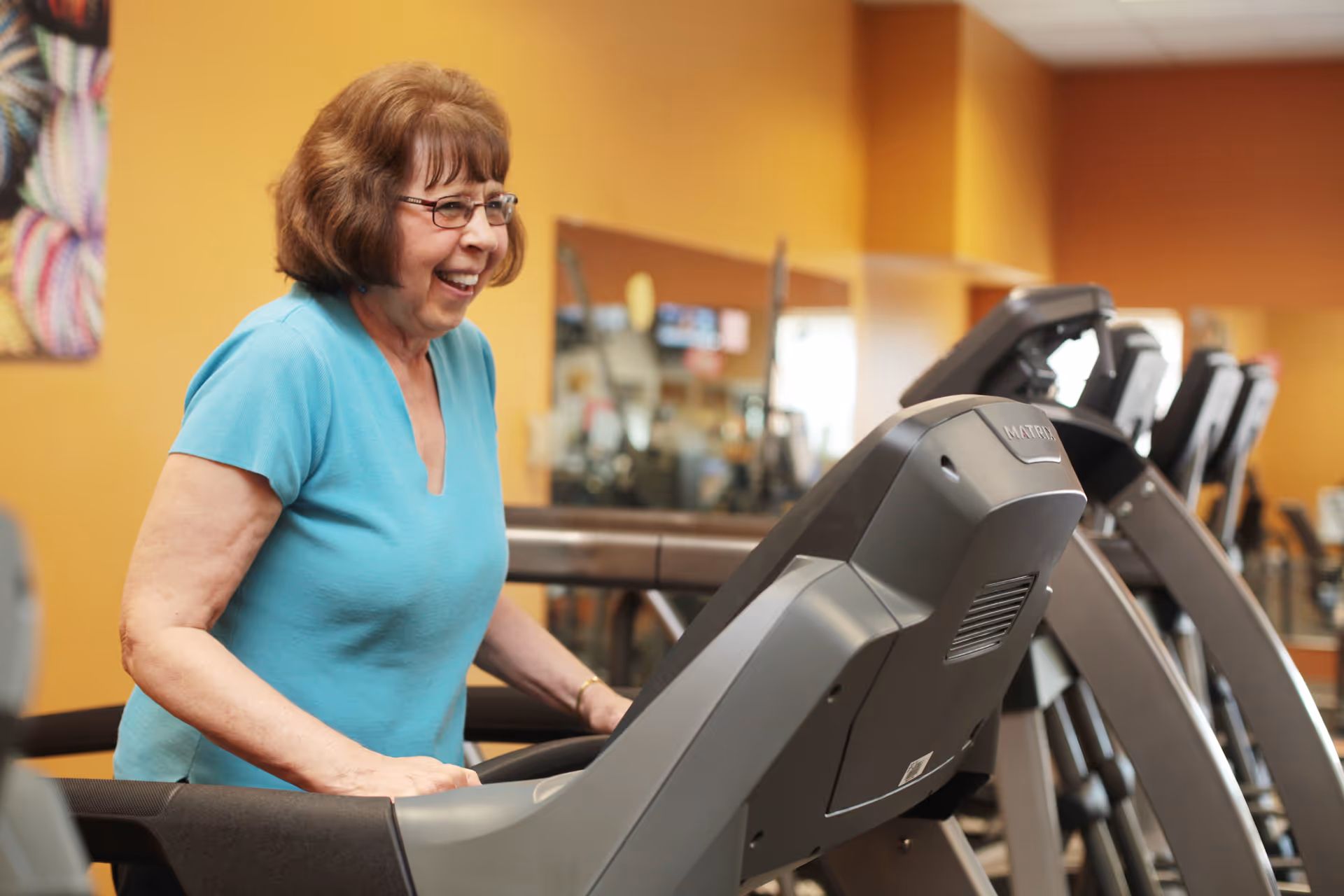 An elderly woman wearing glasses and a turquoise shirt is smiling and walking on a treadmill in a fitness room with yellow walls and exercise equipment in the background.