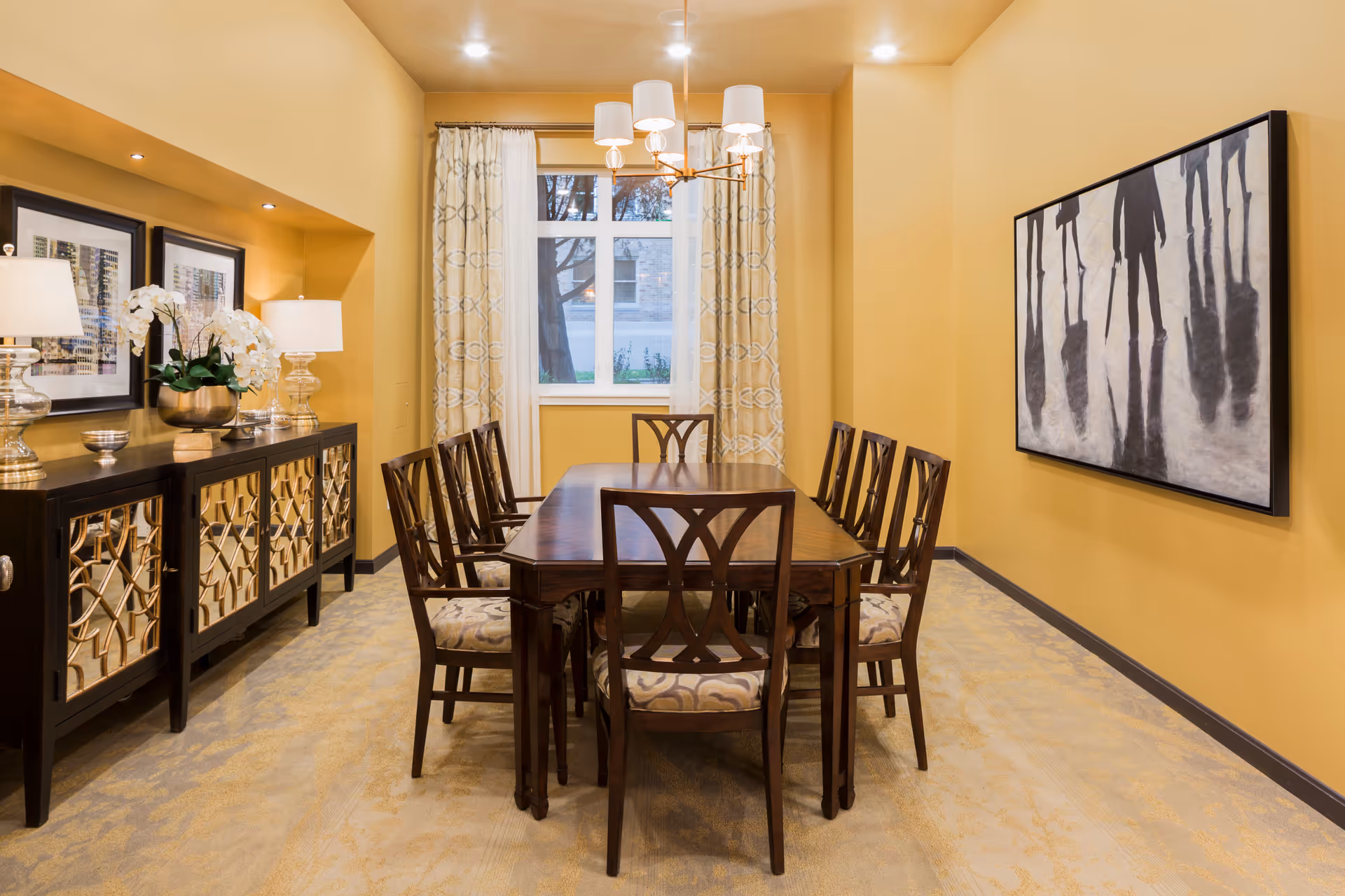 A dining room with a long wooden table surrounded by eight chairs with patterned cushions. The walls are painted yellow, and there is a large window with patterned curtains at the far end. On the left side, there is a dark wooden sideboard with decorative lamps, a plant, and framed artwork above it. On the right wall, there is a large black and white painting depicting silhouettes of people.