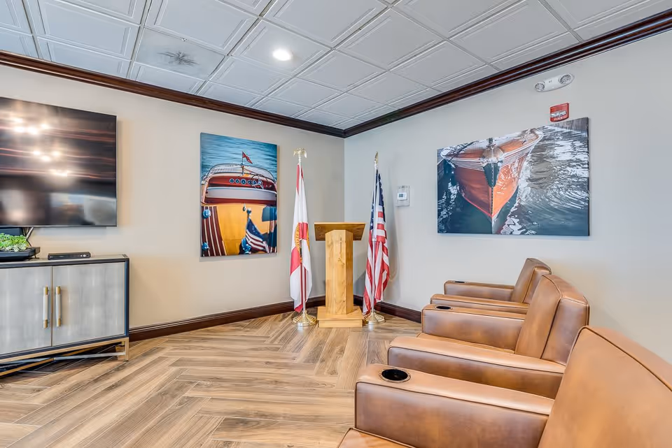 A senior living facility common area with four brown leather chairs arranged in a row, each with a cup holder. On the wall behind the chairs are two large framed pictures of boats on water. Between the pictures are two flags and a wooden podium. To the left is a cabinet with a flat-screen TV mounted above it. The floor has a wood-patterned tile design and the ceiling has a white decorative tile pattern.