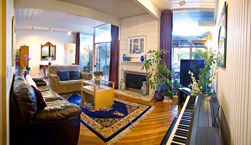 A cozy living room with wooden floors, a dark leather sofa, a beige loveseat, a glass coffee table on a decorative blue and beige rug, a fireplace with vases on the hearth, a piano with flowers on top, and large windows with purple curtains letting in natural light.