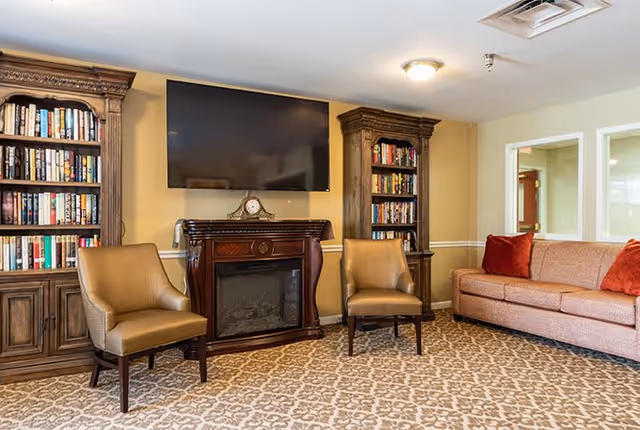 A cozy living room area with a patterned carpet, two brown armchairs, a pink sofa with red cushions, two wooden bookshelves filled with books, a dark wooden electric fireplace, and a large flat-screen TV mounted on a beige wall.