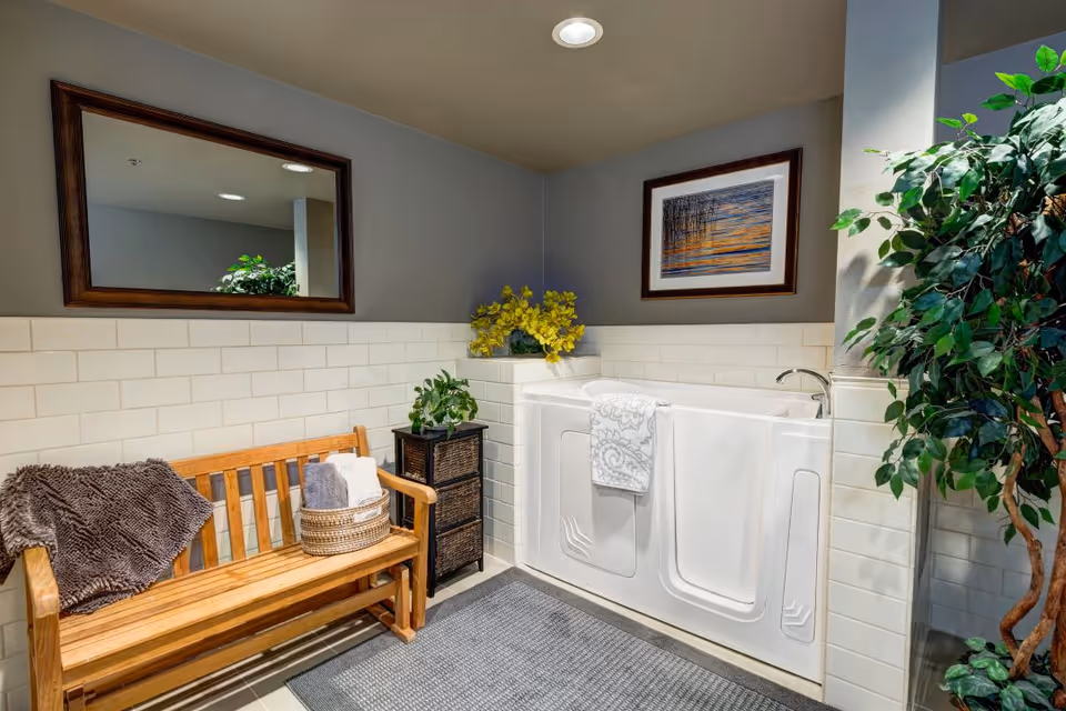 A walk-in bathtub in a bathroom setting with white tiled walls and a gray floor mat. There is a wooden bench with a brown blanket and a basket of towels on it. A small black cabinet with a green plant on top is next to the bathtub. A framed picture hangs on the wall above the bathtub, and a large mirror with a wooden frame is mounted on the adjacent wall. A tall green plant is positioned in the corner.