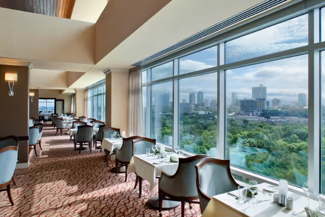 A bright dining room with large floor-to-ceiling windows offering a panoramic view of a city skyline and green trees. The room features tables covered with white tablecloths, set with glassware and napkins, and surrounded by upholstered chairs. The carpet has a red and beige pattern, and the ceiling has wooden accents with modern wall sconces providing lighting.