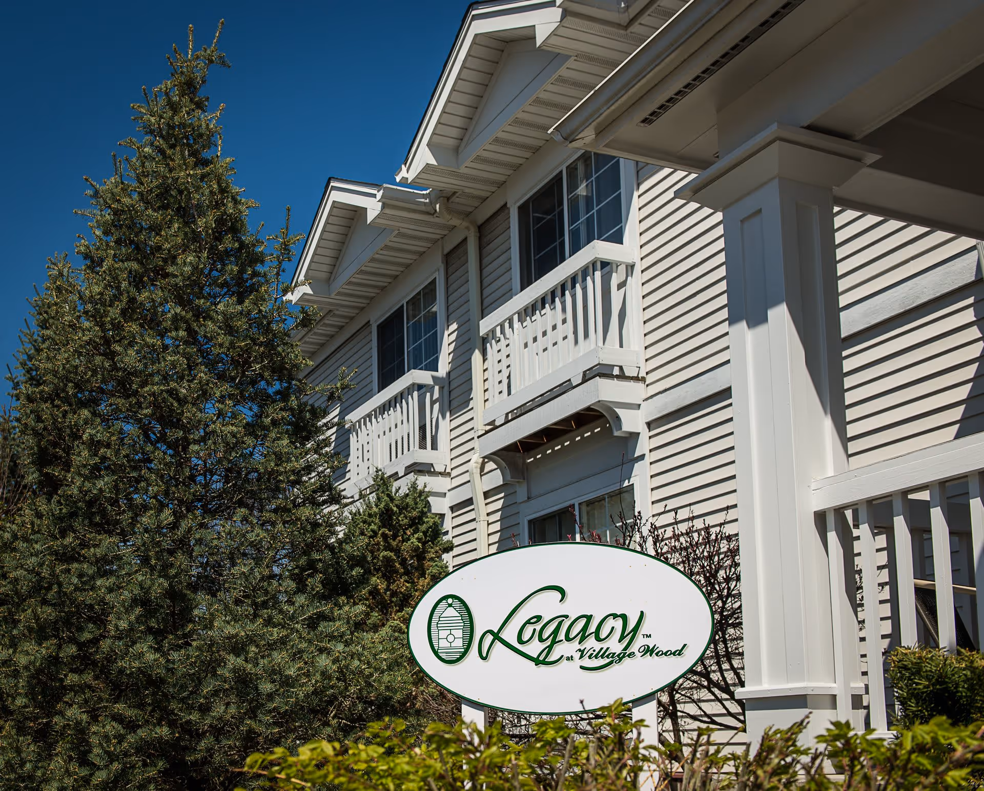 Exterior view of a light-colored building with white trim and small balconies. In front of the building is a sign that reads 'Legacy at Village Wood' surrounded by greenery and trees under a clear blue sky.