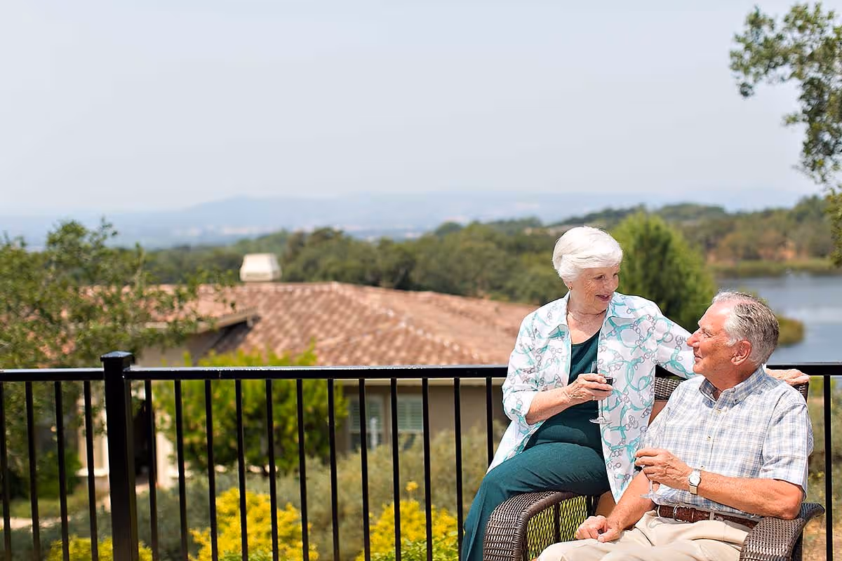 An elderly woman and man sitting outdoors on a patio with a black metal railing, enjoying drinks and smiling at each other. Behind them are trees, a building with a tiled roof, and a body of water in the distance under a clear sky.