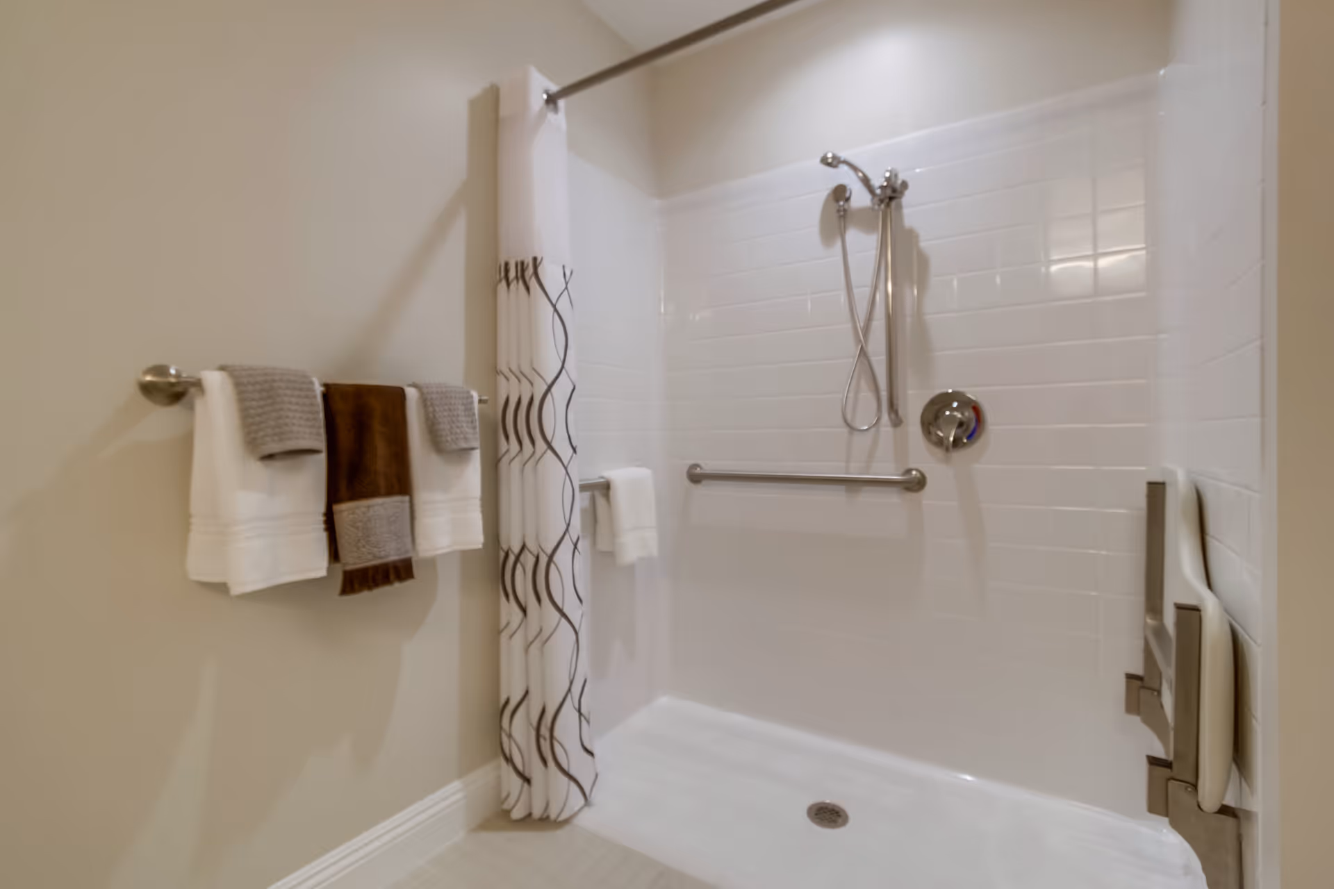 A clean, accessible shower area with white tiled walls, a handheld showerhead, a horizontal grab bar, and a foldable shower seat mounted on the right wall. A shower curtain with a white and brown wave pattern is partially drawn. On the left wall, there is a towel rack holding three towels in white, brown, and gray colors.