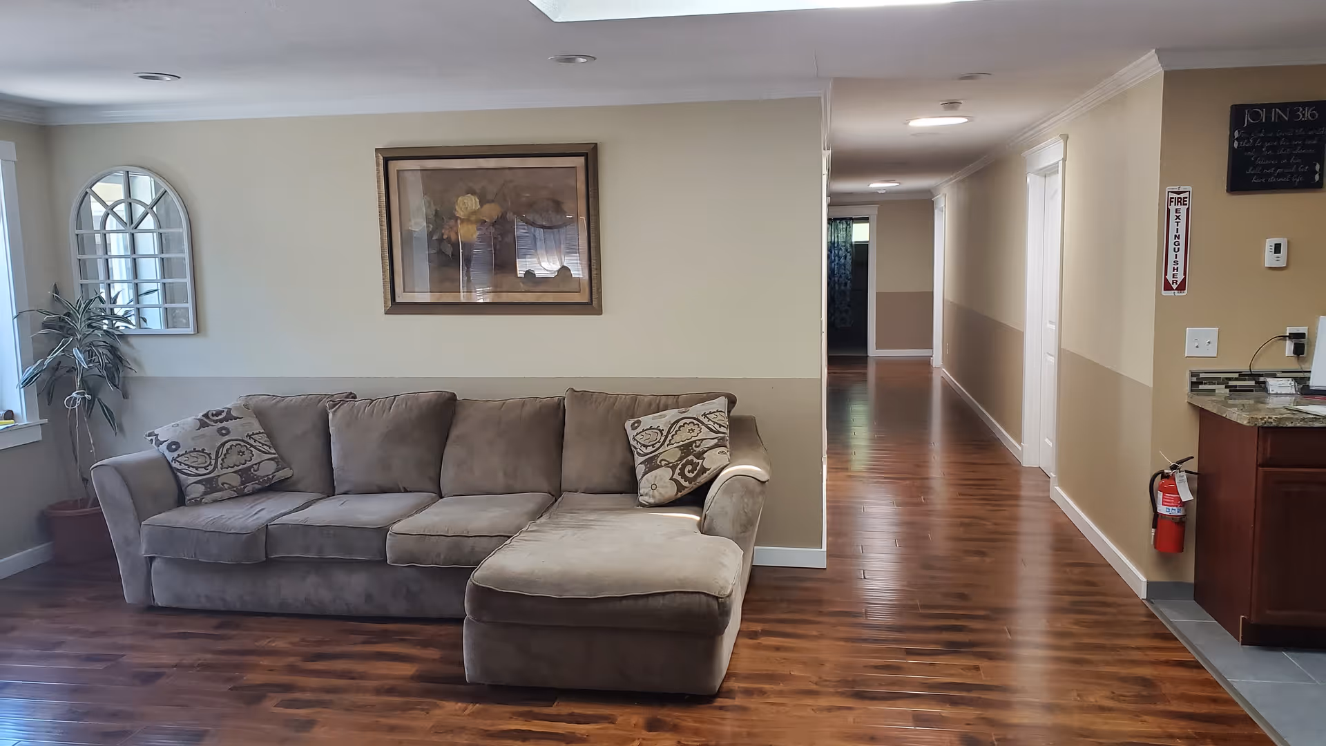 A living room area with a beige sectional sofa adorned with two patterned pillows. The room has wooden flooring, a potted plant near a window with white blinds, a decorative wall mirror, and a framed floral painting above the sofa. To the right, there is a hallway leading to other rooms and a section of a kitchen counter with a fire extinguisher mounted on the wall.
