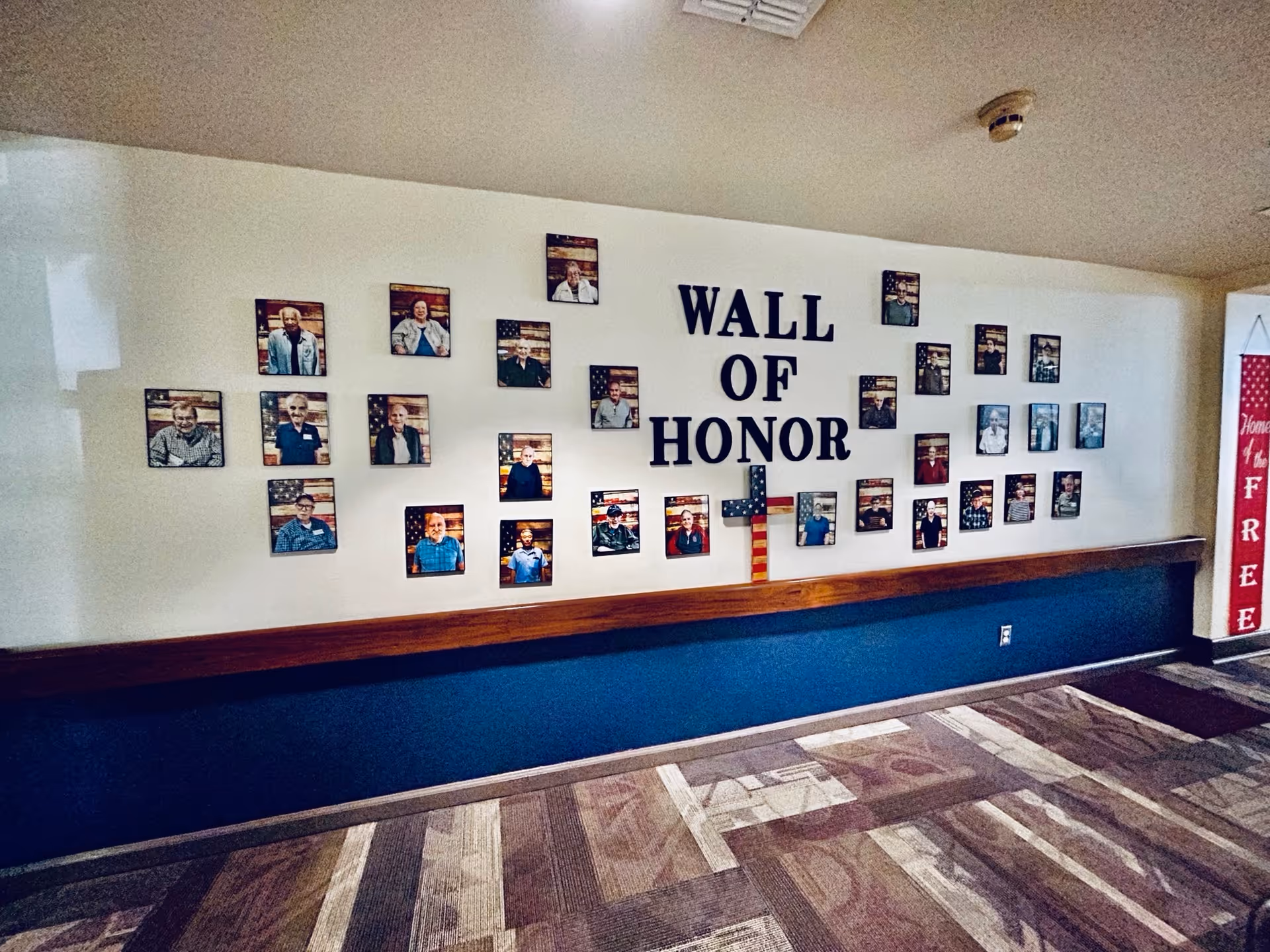 Interior hallway showing a 'Wall of Honor' display of framed portraits and an American-flag cross on a white wall above a blue wainscot.