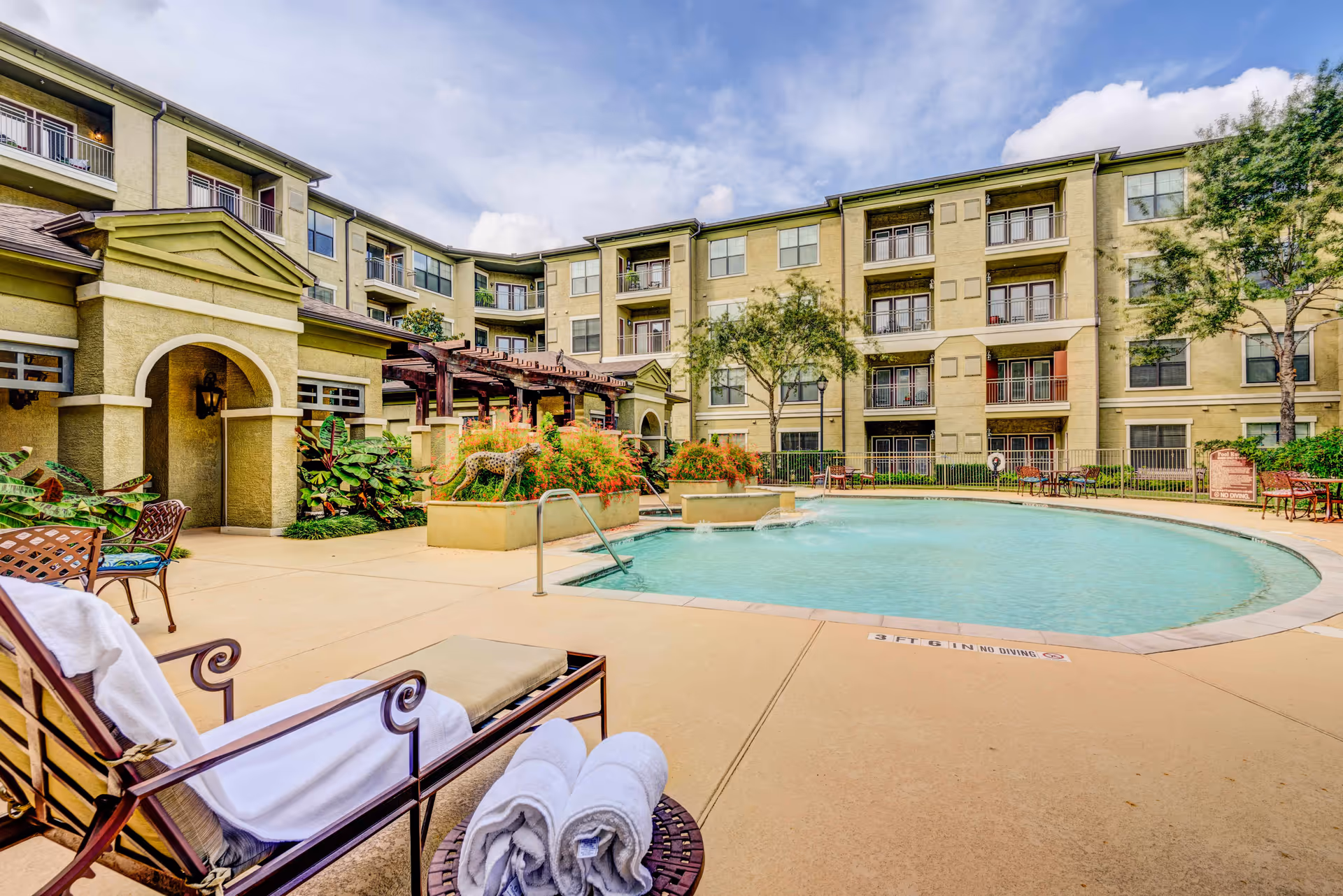 Outdoor swimming pool area at Conservatory At Champion Forest with lounge chairs, rolled towels, and a multi-story residential building in the background under a partly cloudy sky.