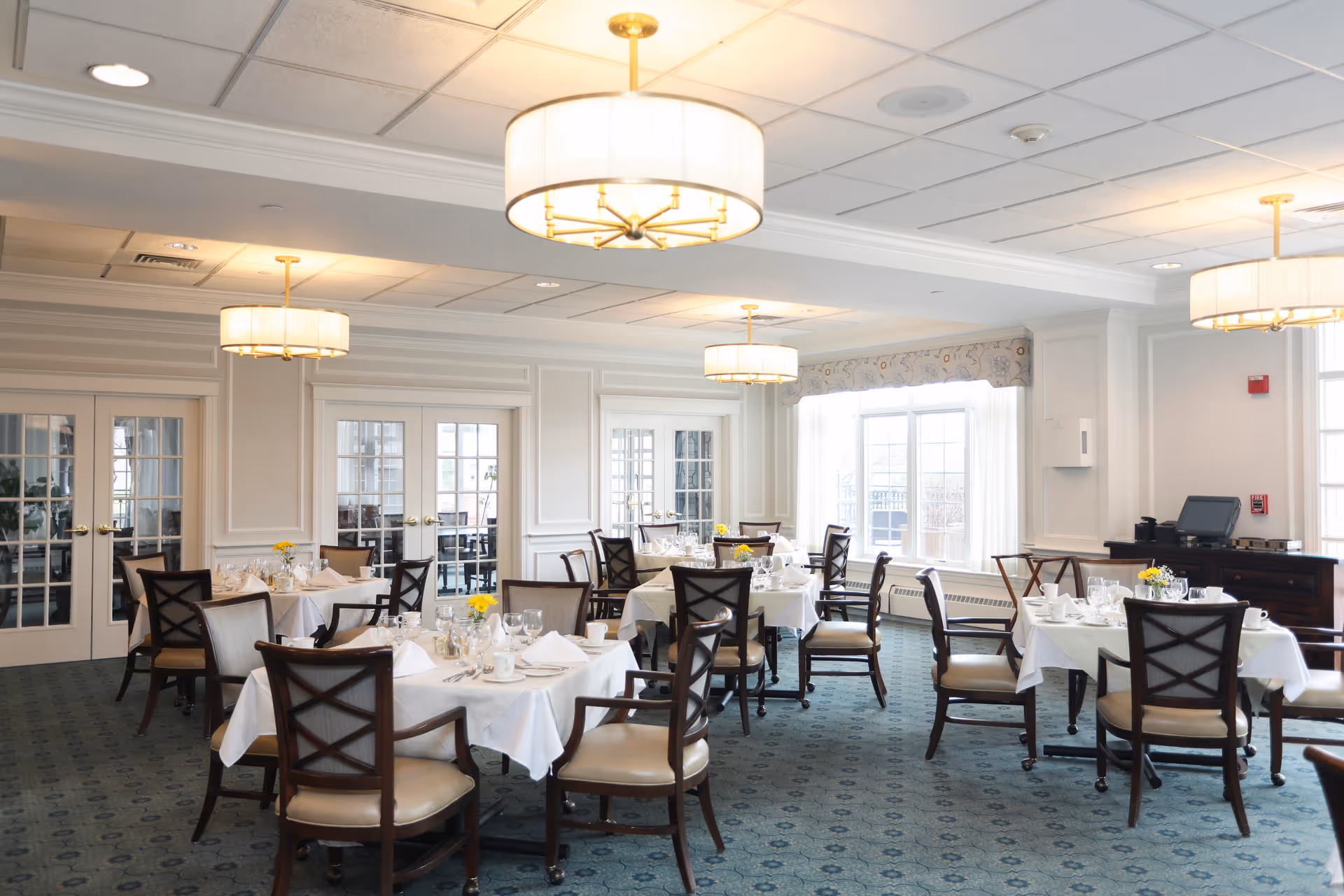 Bright dining room with neatly set tables, cushioned chairs, large windows, and drum chandeliers.