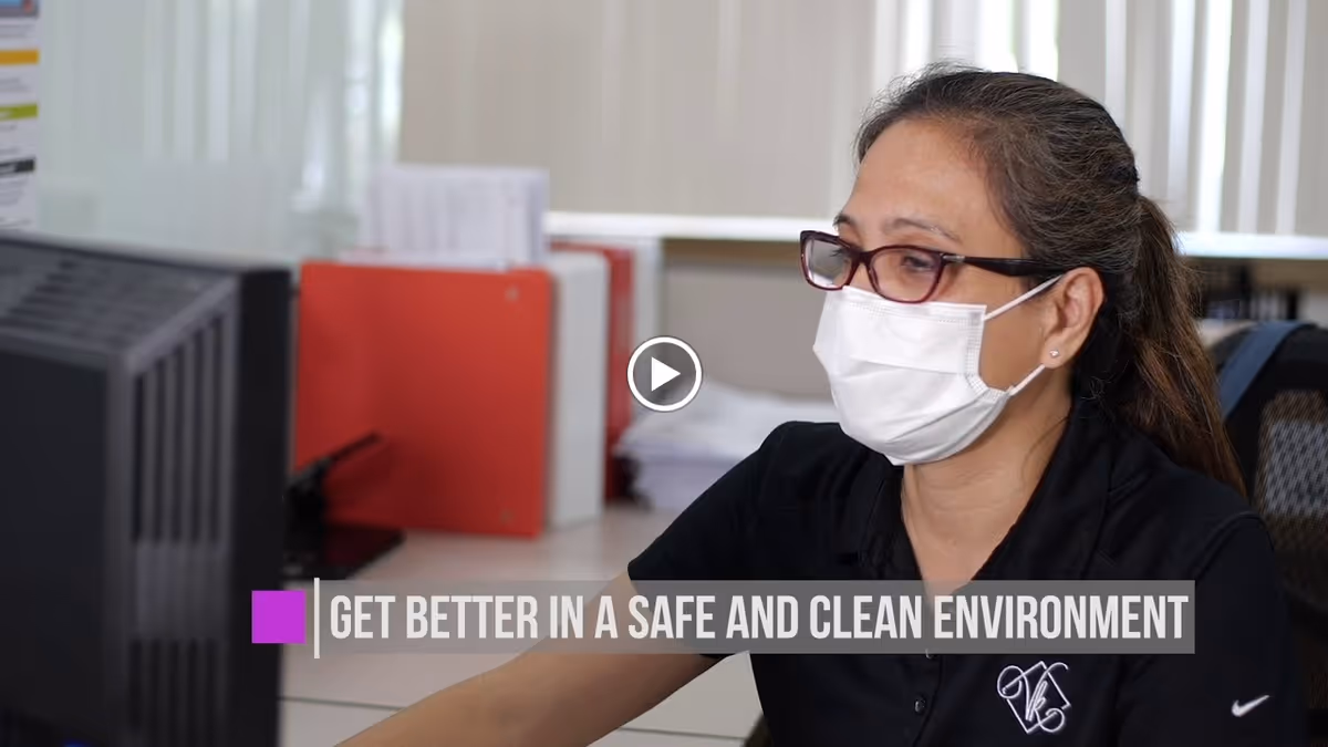 A woman wearing glasses and a face mask is sitting at a desk, looking at a computer monitor. Behind her are office supplies including red binders and papers. Text on the image reads 'GET BETTER IN A SAFE AND CLEAN ENVIRONMENT.'