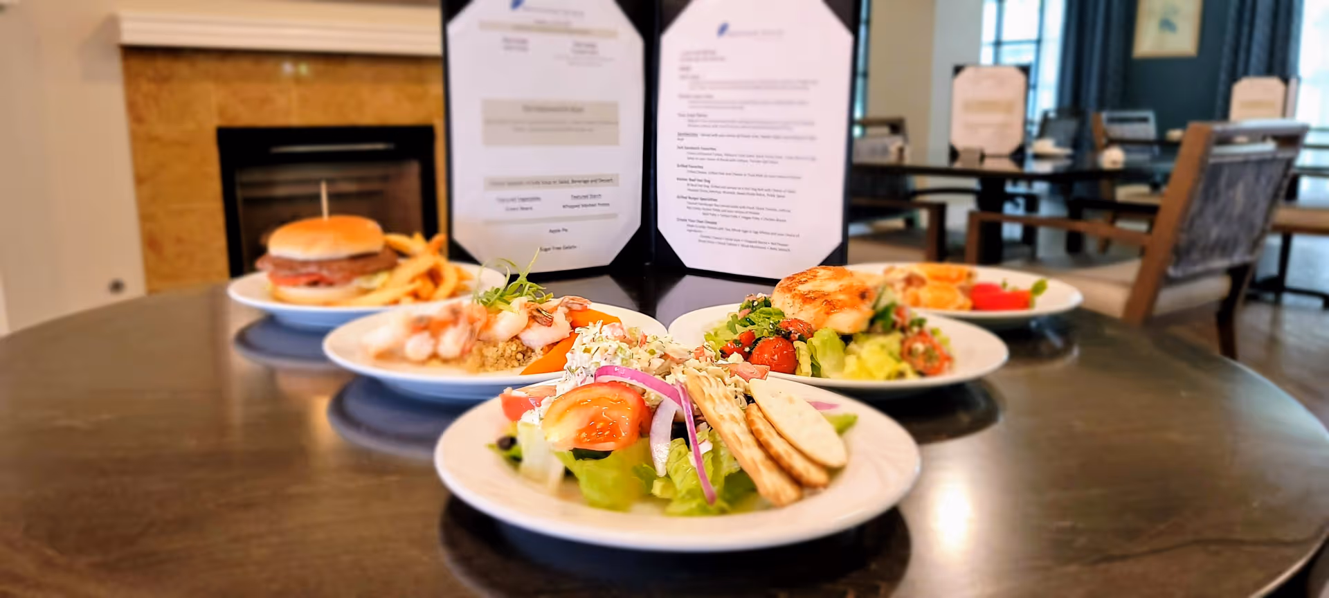 Several plated meals including salads, shrimp, and a burger on a round dining table with menus and chairs in the background.